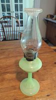 Full view of antique Mosser Glass jadeite green oil lamp with clear glass chimney, showing the entire lamp on a wooden table with background chair and window.