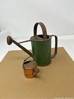 Green antique watering can with curved spout and arched handle alongside a small copper miniature watering can, placed on brown surface and photographed from side angle.