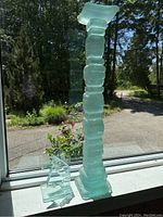 Tall greenish translucent glass column sculpture displayed on window ledge outdoors.
