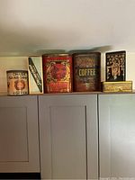 Five vintage tins displayed on a shelf including coffee and tea tins from Chase Sanborn, Smallman Campbell, Lipton, W.H. Schwartz & Sons, and an Asian-themed decorative tin. All show significant wear and vintage patina.