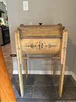 Front view of yellow wooden churn with black painted 'B' emblem and decorative floral stencils on barrel, showing four legs and metal hand crank on side.