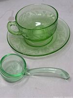 Green translucent teacup and matching saucer with shamrock/clover etched pattern, accompanied by a small green glass dipper spoon placed beside the set.