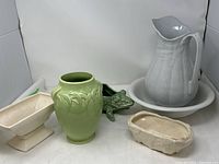 Five vintage McCoy pottery pieces arranged in a corner: white pitcher and bowl, two ivory planters, green leaf patterned vase, and frog planter