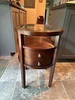 Front view photo showing the round wooden side table with beveled glass top, one of the drawers partially visible with silver knob handles.