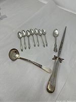 Full view of all items: 7 mini spoons, 1 tasting spoon with mother of pearl handle, 1 sterling teaspoon, 1 meat poker with broad guard and pointed tip.