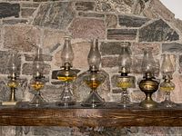 Seven vintage oil lamps arranged in a row on a wooden shelf in front of a stone wall. Two lamps have bronze bases, others have clear glass bases. Lamps have glass chimneys and are partially filled with yellowish oil.