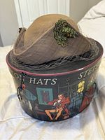 Brown ladies hat with green applique and net veil sitting on top of original Stetson decorative round hat box with vintage illustrated pattern