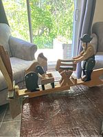 Side angled view of handcrafted wooden whirligig with detailed figures of man cutting wood and woman washing clothes. Visible windmill blade on left side.