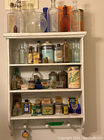 View of full white wooden medicine cabinet filled with vintage and antique clear and colored embossed glass bottles, collectible medical tins, and glass eye wash cups