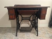 Front view of Singer sewing machine cabinet showing wrought iron base with the brand name, wooden top, and two side compartments.