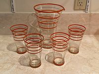Photo showing one pitcher and four glasses with concentric orange and gold lines on clear glass on a countertop.