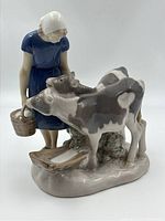 Front side showing woman in blue dress with white bonnet standing with two black and white cows near feeding trough, holding bucket.