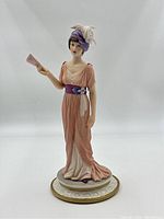 Front view of porcelain figurine showing details of dress, fan in right hand, and feathered headband.