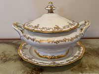 Front view of the soup tureen set on a reflective surface showing gold floral decoration and handles