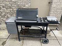 View of Weber natural gas grill showing front, including lid with thermometer, control knobs, side burner with cover, side prep area, lower wire shelf, and attached gas hose.