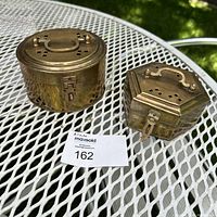 Top view of two vintage brass cricket boxes; one round and one hexagonal with lids closed showing vents and handles.