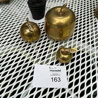 Three brass apple items arranged on a white metal mesh table. The items include two smaller apple bells and one large apple box with a removable lid.