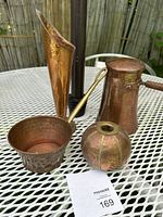 Four copper items displayed on outdoor table: a tall hammered copper bud vase, a small hammered copper pot with brass handle, a copper candleholder, and an antique copper lidded chocolate pot with wooden handle.