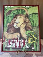 Front view of framed artwork showing the lion and turtle playing chess with red and white pieces, under red wooden frame.