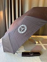 Black umbrella opened displaying Mercedes-Benz emblem on canopy alongside matching sleeve on table surface.