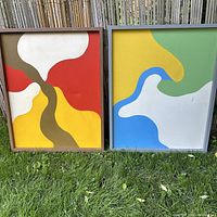 Photo of both vintage abstract mod paintings outdoors on grass in front of a wooden fence, showing colorful abstract designs with visible dents and scratches on canvas and frames.