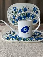 Photo showing white metal watering can with blue flower design placed on two vinyl placemats with blue floral patterns, highlighting the set.