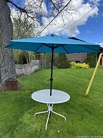 Photo of entire patio table with turquoise umbrella open outdoors on grass with some wear visible on the table's white paint.