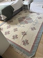 Wool rug laid out on floor, showing full central floral pattern and surrounding geometric crosses in pastel tones with visible fringe on one edge.