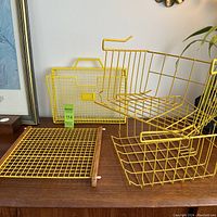 Photo showing all items from the lot: two yellow wire hanging baskets, a yellow wire trivet or wall rack with wooden frame sides, and a yellow wire wall folder or mail holder on a wooden surface.