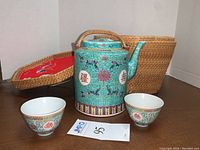 Woven wicker picnic caddy with teal colored Asian tea pot and two cups, showing intricate floral and Chinese character designs.