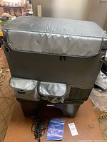 Front view of gray insulated cover with two small front pockets, sitting on black Hubert stand with cords and manual in front.