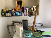 Photo of assorted cleaning products on shelf including Husky contractor cleanup bags, spray cleaners, and other bottles with some items on countertop including black tower fan heater and white dehumidifier.