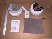 Two wireless eye massagers, one white and gray Renpho with USB cable and packaging, the other white and black Mya with a drawstring pouch and a ruler for scale
