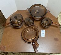 Full set of six brown translucent glass Corning Visions cookware items including lids and handles arranged on table.