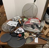 Full shot of all items arranged on a table showing cast iron skillet, meat press, utensils, grilling baskets, and gas valve hose.
