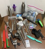 Full overview of kitchen utensils including water bottles, knives, whisks, spatulas, measuring tools, and ladles spread out on a brown surface.