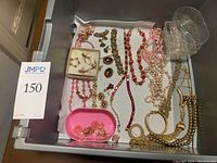 Photo of lot jewelry laid out in storage drawer showing mix of pink, red, and gold costume jewelry including necklaces, earrings, and bracelets.