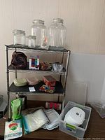 Photo of the full HDX metal shelf fully assembled, with FoodSaver glass jars on top shelf, assorted containers, picnic basket, reusable plastic bags in bin on floor, and other items arranged on shelves and floor.