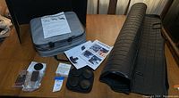 Full view of lot items including grey car cover in bag, Tesla Model X rubber trunk mat rolled, instruction sheets, LED shadow light, and small packaged items on wooden table