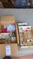 Photo of assorted costume jewelry items laid out in two cardboard boxes on a beige fabric surface. Visible items include various bracelets, earrings on cards, necklaces, brooches, and hair combs.