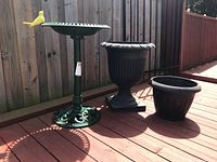 Green metal bird bath standing on wooden deck next to two black plastic planters of different sizes.
