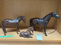 Three hand carved wooden horse figures displayed on shelf including a large horse with leather accessories, medium horse, and a small foal lying down.