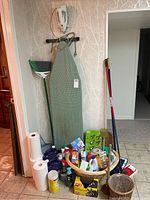 Wide shot of ironing board, brooms, mops, paper towels, and basket with cleaning supplies