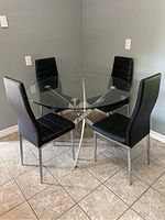 Photo shows round glass table with chrome base and four black leather chairs arranged around it.