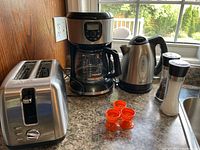 Kitchen countertop with Master Chef 2-slice toaster, Black & Decker coffee maker, Sunbeam electric kettle, salt and pepper grinders, and three orange plastic egg cups grouped together.