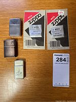 Photo showing three lighters and two unopened Zippo wick packages on a wooden surface.