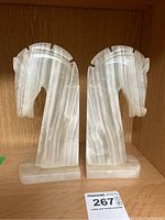 Front view of two white and beige striated marble horse head bookends on wooden shelf with auction label.