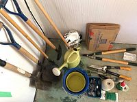 Photo showing four shovels, a green watering can, blue bucket, garden gnome, and some folded cardboard and other items