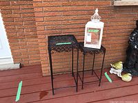 Two black metal nesting tables with intricate lattice tops placed outdoors against brick wall, one slightly smaller and fitting under the other.