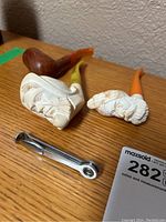 Two hand-carved meerschaum face pipes with yellow and orange stems, a wooden pipe, and a metal pipe cleaner tool on a wooden desk